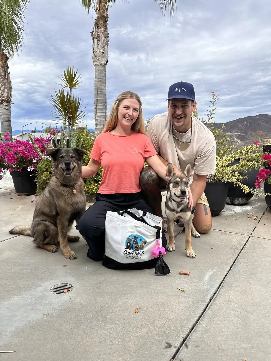 Fawn, a tan and black puppy with oversized ears, standing beside her new family on a patio between potted bougainvillea, a smiling woman in a coral shirt and a smiling man in a navy cap, with the family's other dog and a Comeback Pack tote bag