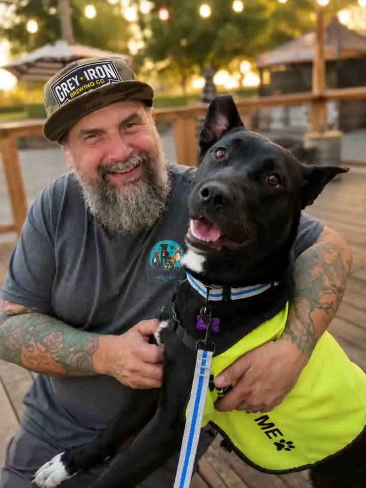 A Comeback Pack volunteer in a Grey Iron Brewing hat smiling while holding a black foster dog wearing a yellow "foster me" vest at the Foster Pup Social