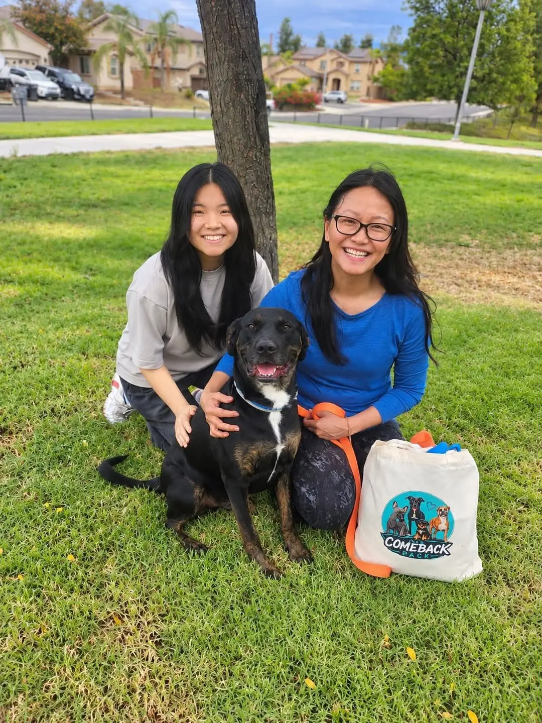 Rickey Henderson, a black and brown dog with a white chest patch, grinning on the grass at a park between a smiling teenage girl and a smiling woman holding his orange leash, a Comeback Pack tote bag beside them