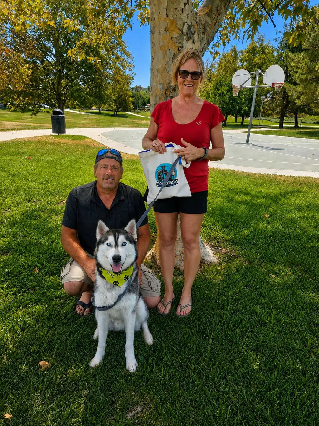 Samson, a black and white husky in a green paw-print bandana, sitting on the grass at a park on send-off day, with a man kneeling beside him and a woman holding his leash and a Comeback Pack tote bag