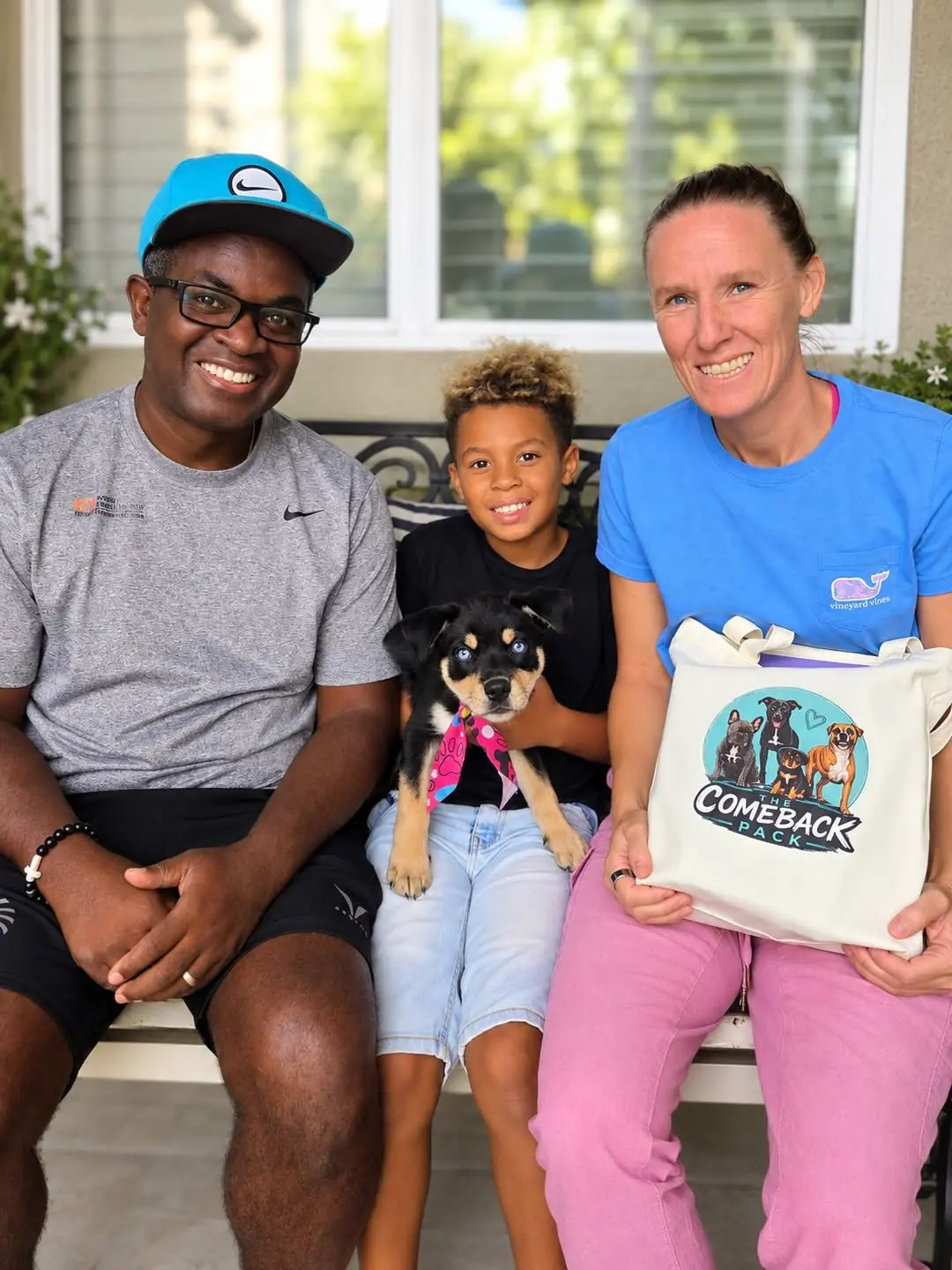 Stormy Skye, a black and tan puppy with blue eyes in a pink paw-print bandana, held on a boy's lap on a porch bench between a smiling man in a blue cap and a smiling woman holding a Comeback Pack tote bag